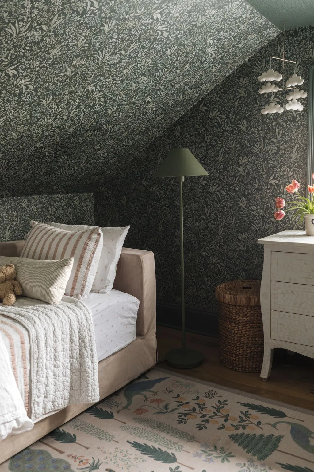 Attic bedroom nook with dark gray-green leaf wallpaper, pale upholstered daybed, white quilt, striped pillow, green floor-lamp shade, and tall basket.