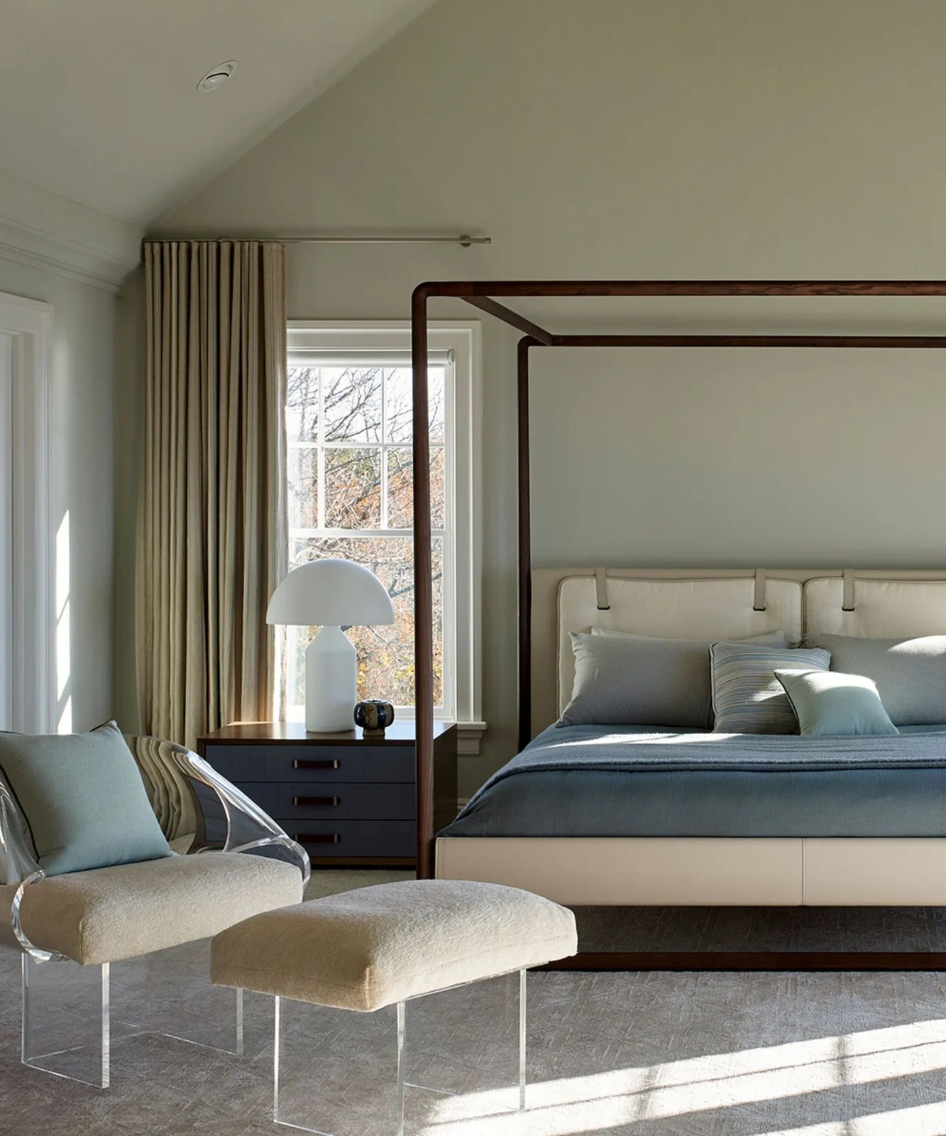 A slim wooden canopy frame surrounds a pale upholstered bed with blue bedding beside a light chair, ottoman, white lamp and tall curtains