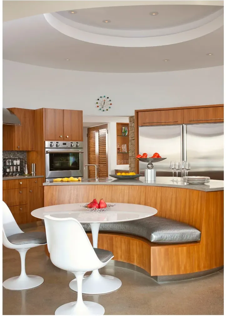 A curved wood banquette around a white pedestal table with white chairs, beside a wood kitchen and colorful clock.