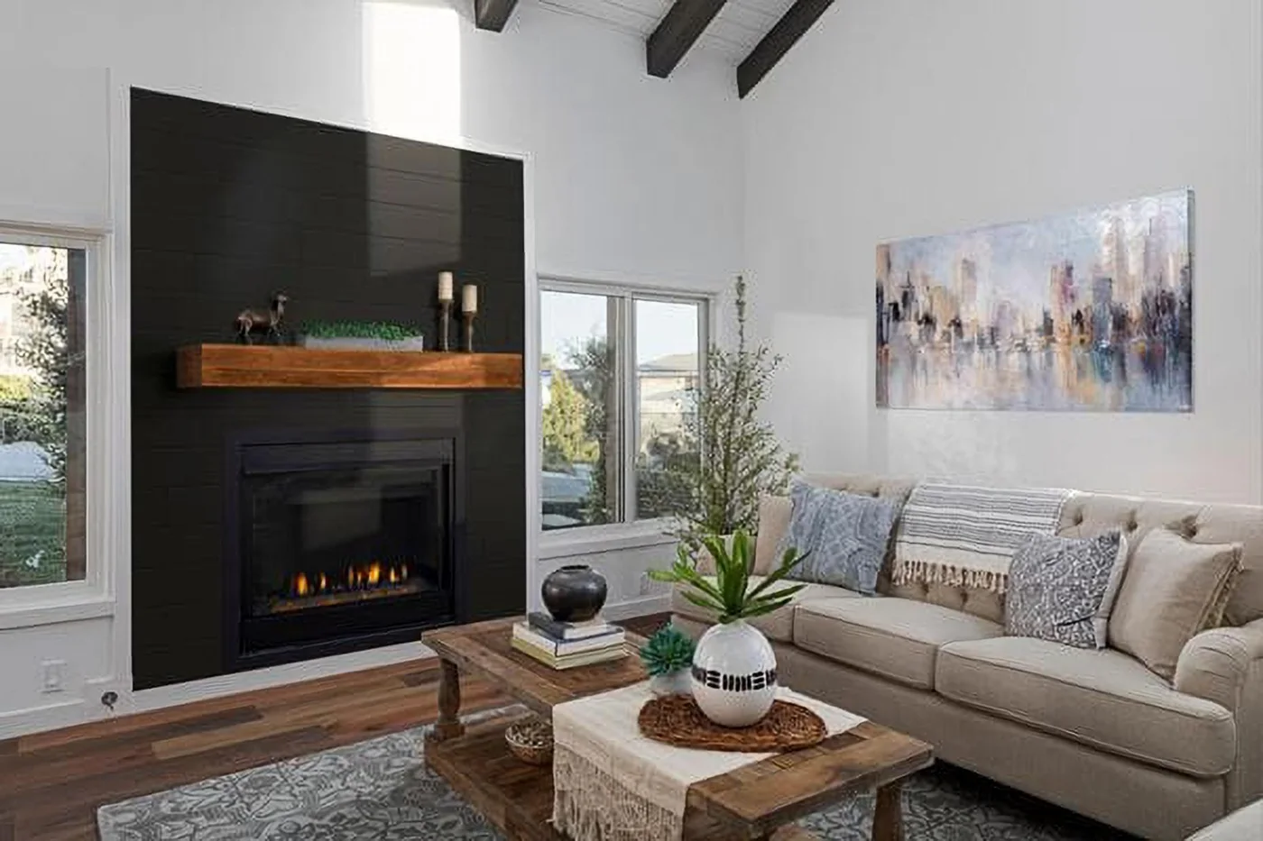 A black horizontal shiplap fireplace wall has a wood mantel and dark insert between windows, beside a pale sofa, wood tables and patterned rug.