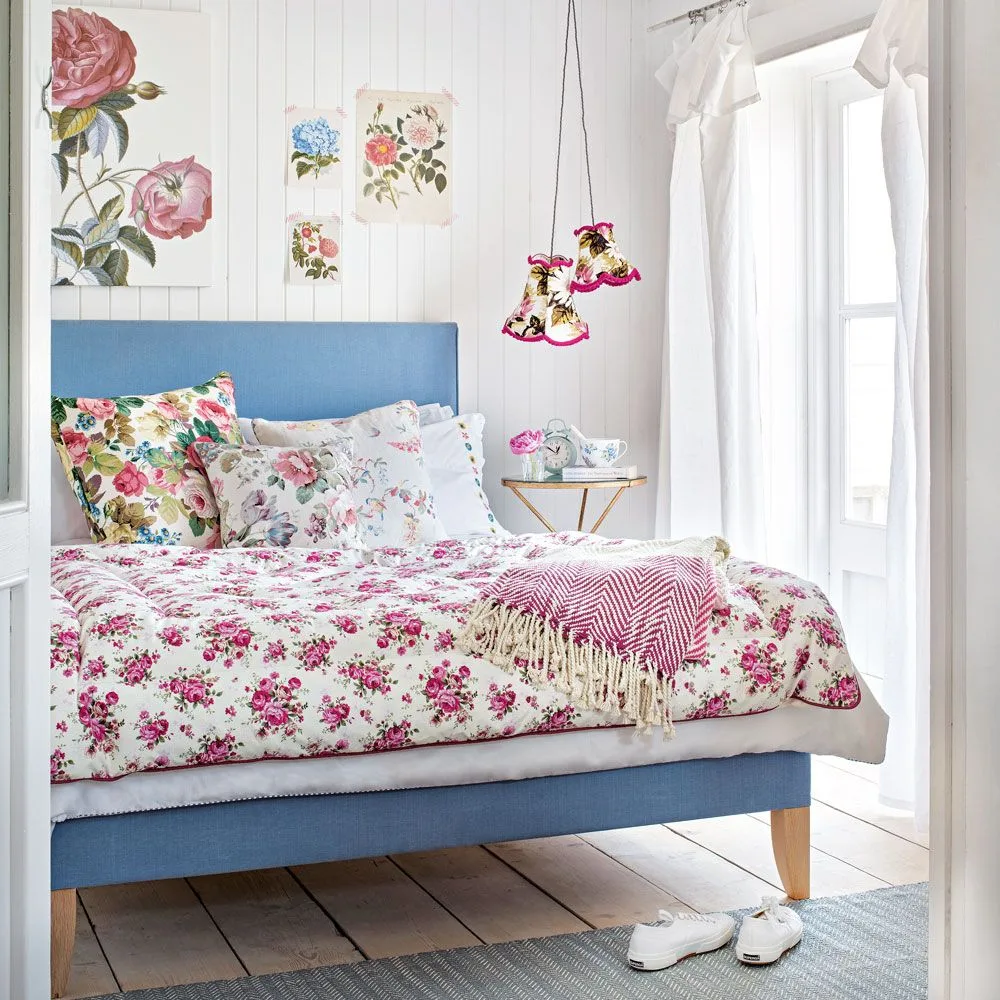 A blue upholstered bed stands against white vertical boards, with mixed floral bedding, botanical prints, small hanging shades and a narrow bedside table.