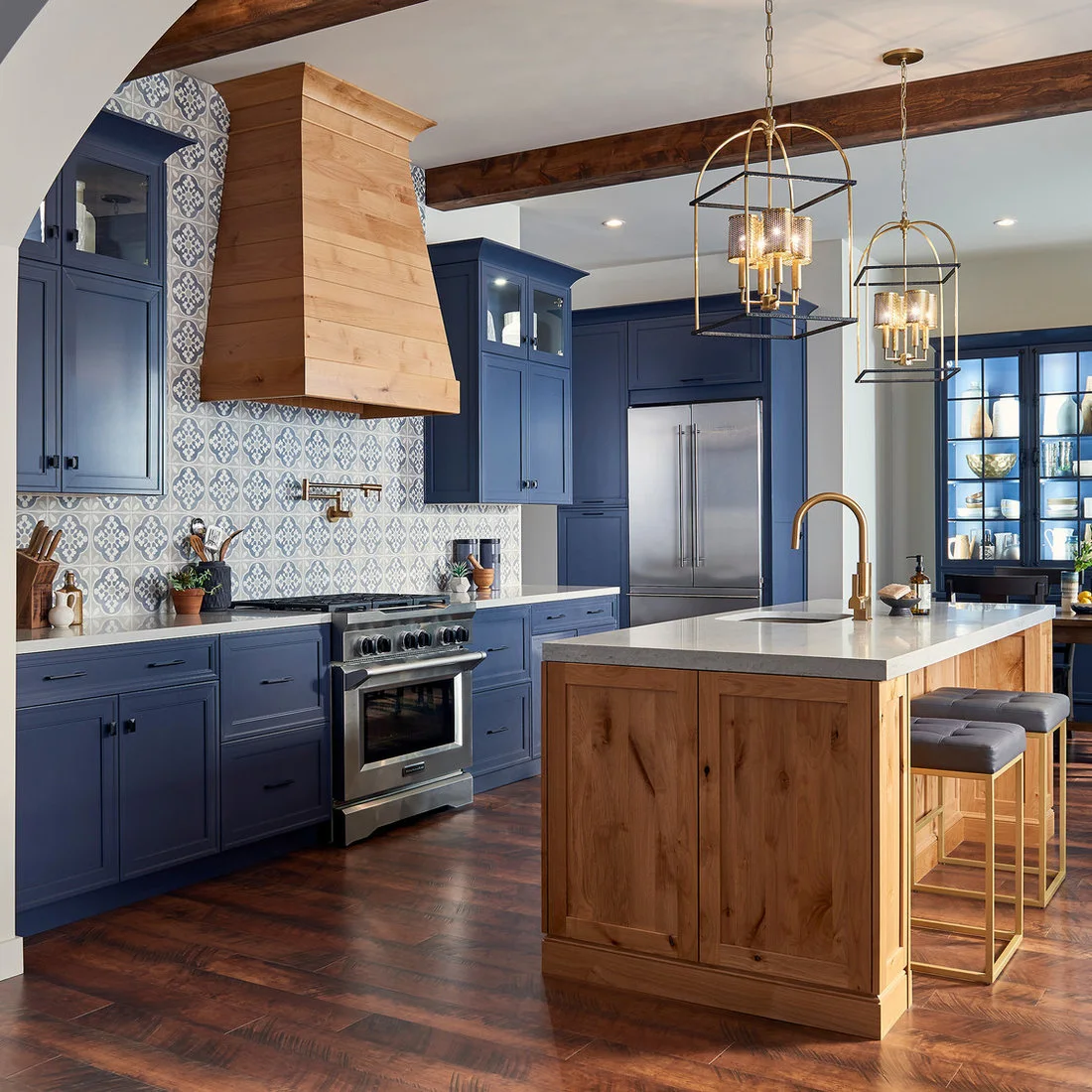 Blue cabinetry and patterned tiles surround a timber island beneath ceiling beams and lantern pendants.