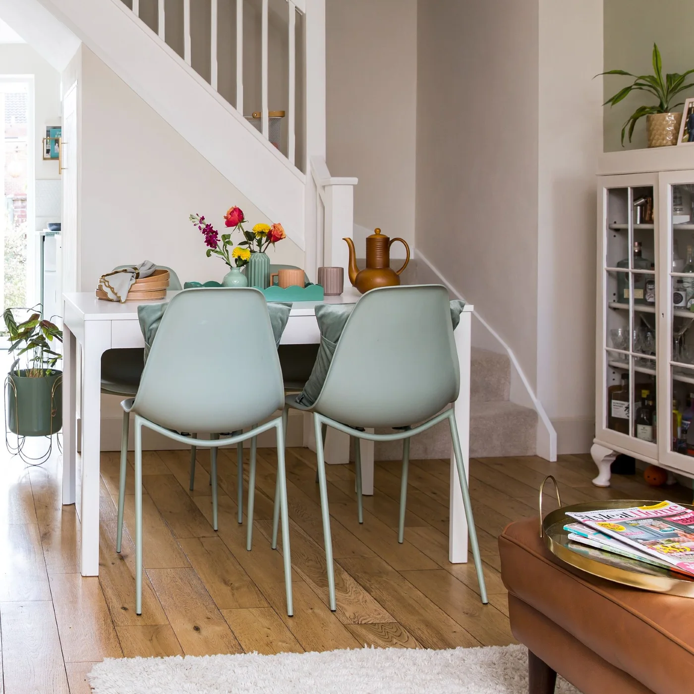A small white dining table with mint chairs stands against a stair wall beside a glazed cabinet and living-room seating.
