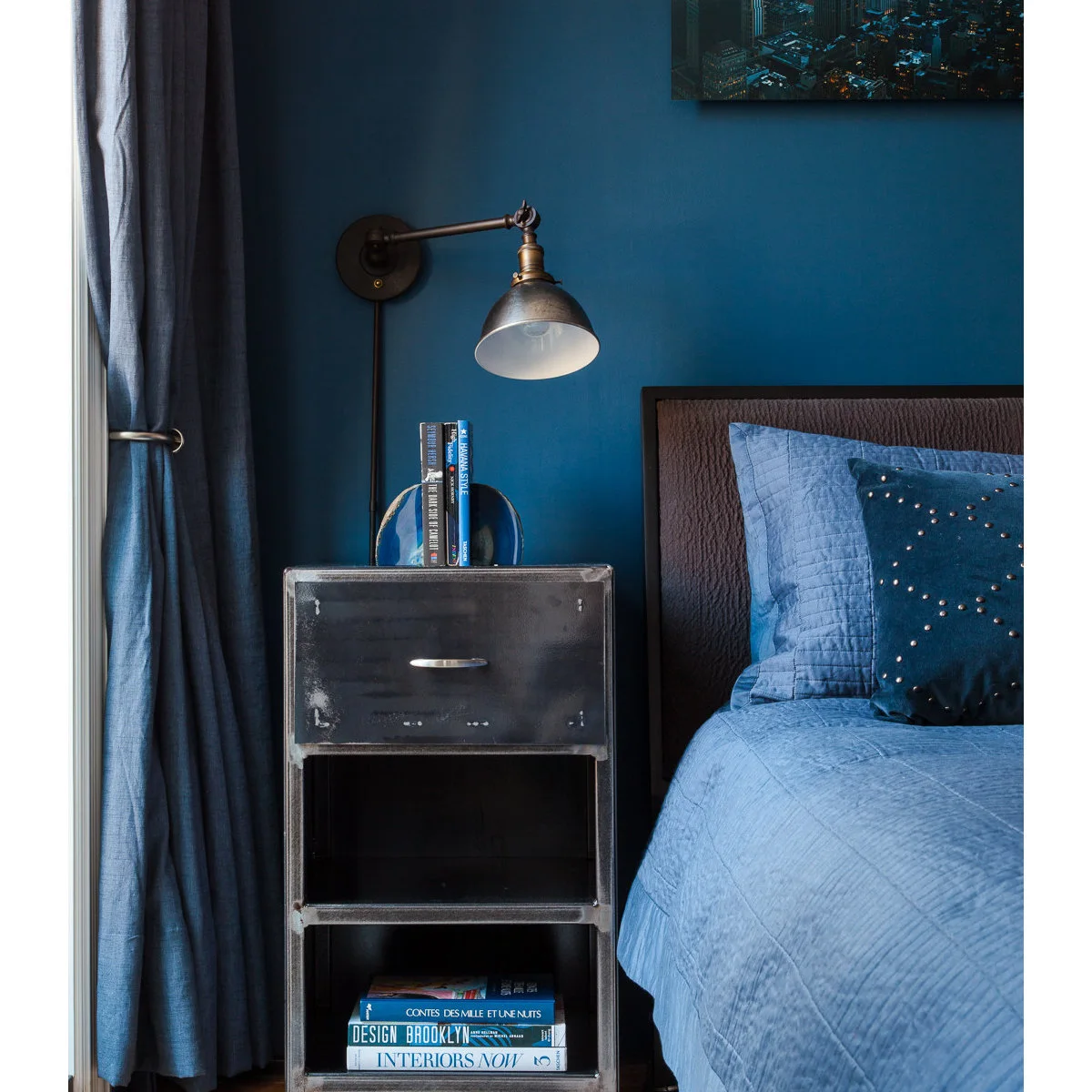 Blue bedroom with worn metal-looking bedside cabinet, books and an adjustable wall light
