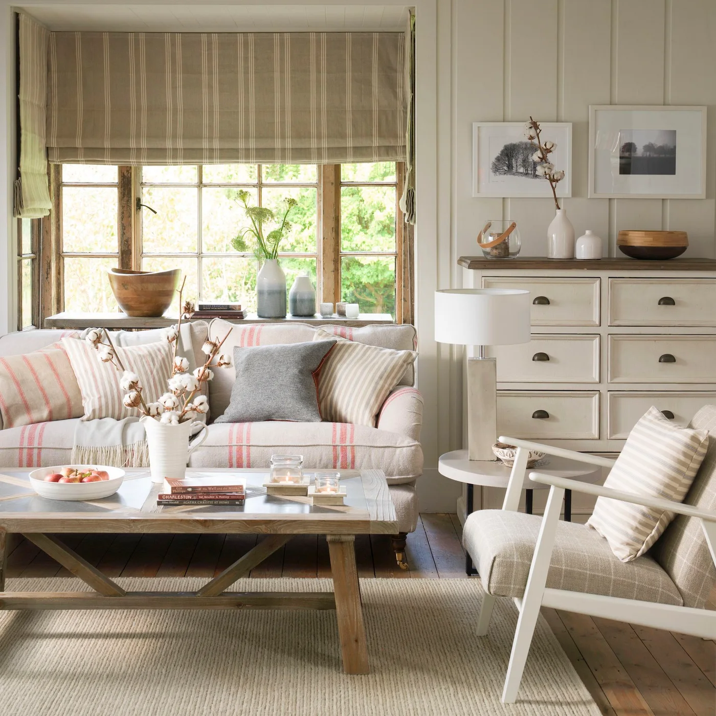 A pale striped sofa and white-framed chair surround a wood coffee table beside painted drawers and a timber-framed window.