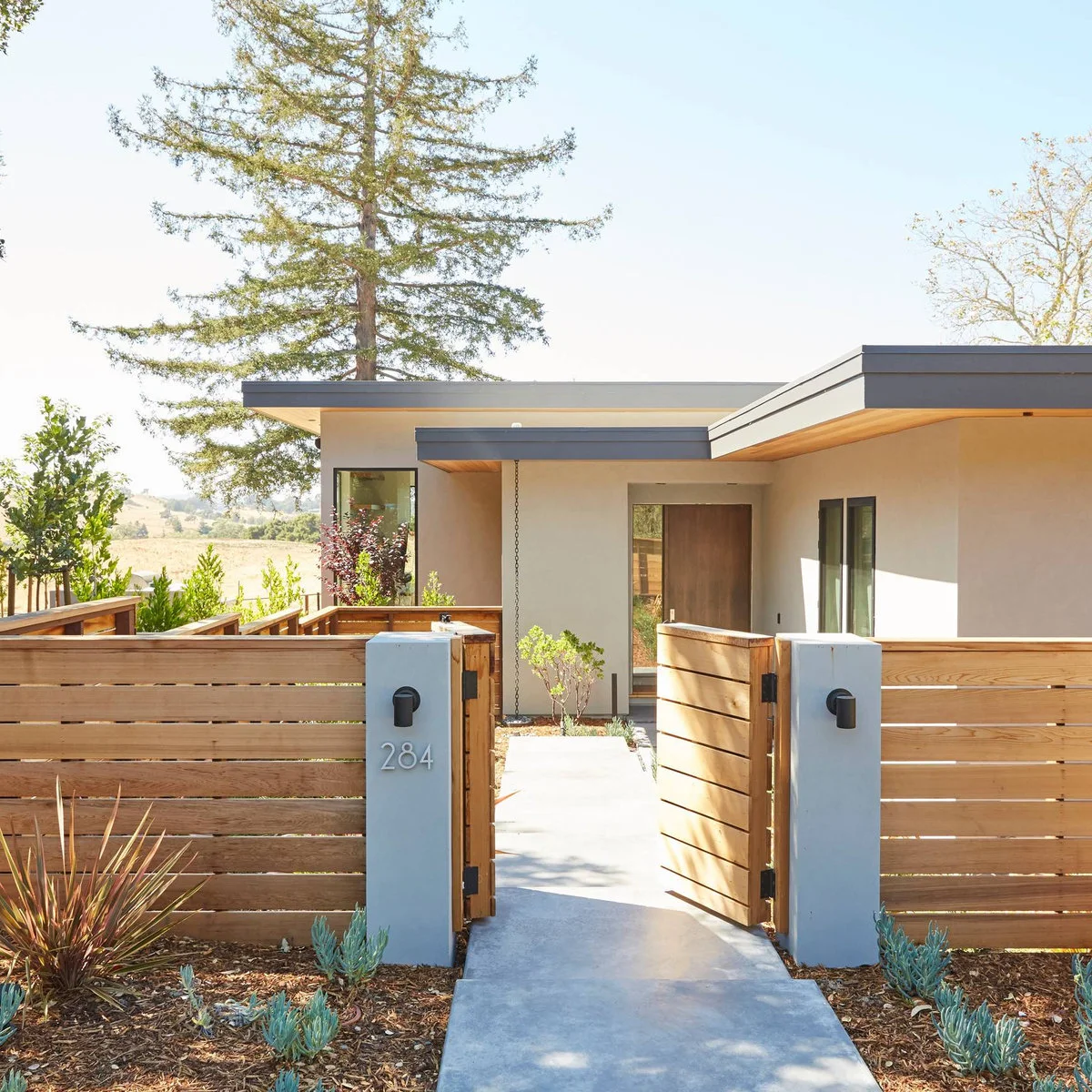 Horizontal wood fence boards meet pale solid gate posts with small lights and house numbers beside an entrance path.