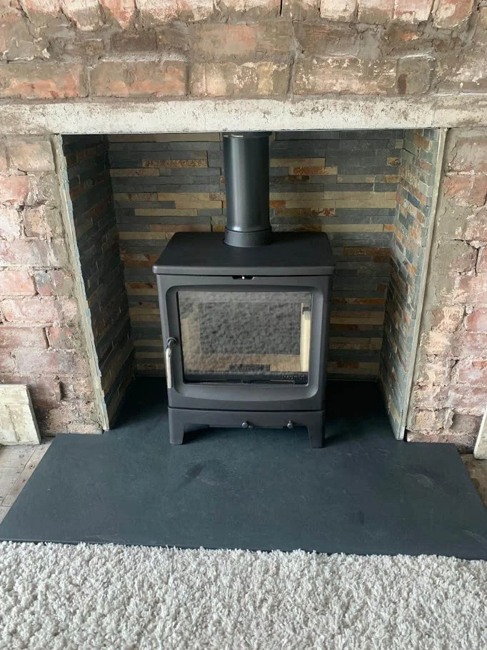 A black stove stands within a rough brick opening above a dark slate hearth with a broad projecting front.