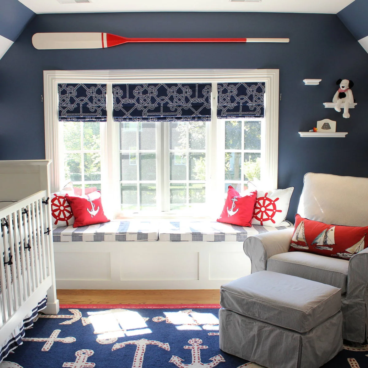 Navy nursery with a striped window seat, red nautical cushions and a pale chair with matching ottoman