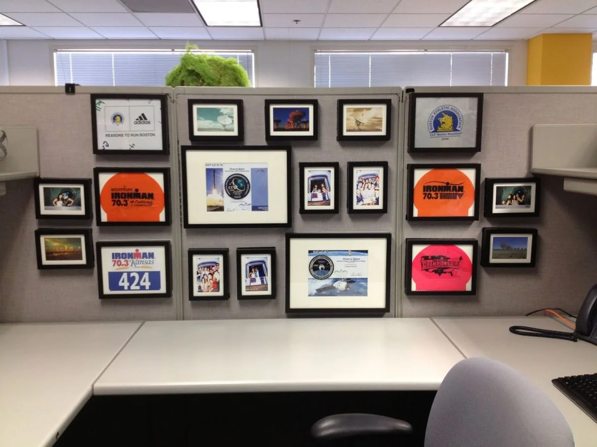 Black frames holding race mementos, photographs and small landscape pictures across gray cubicle panels above a largely clear desk.