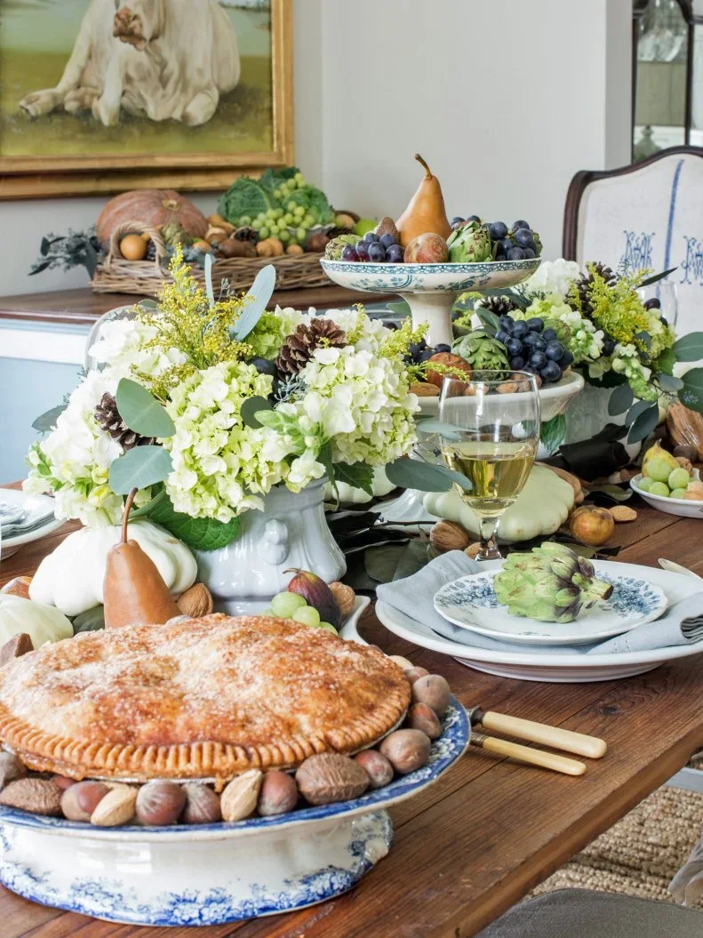 Pale green flower arrangements and fruit bowls sit beside white pumpkins, patterned plates and a pie on a wooden table