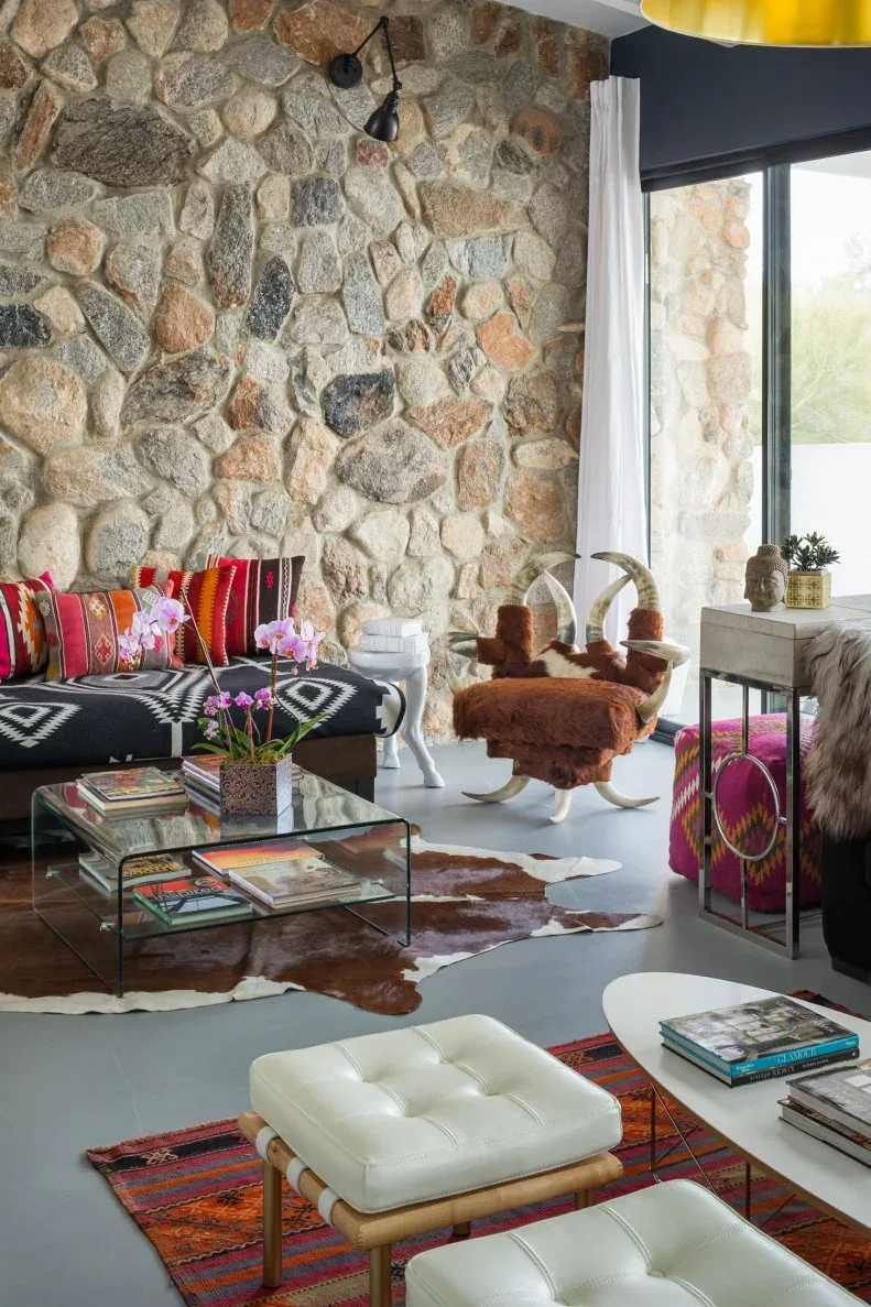 Stone-walled living room with patterned seating, red cushions, a glass coffee table and layered rugs.