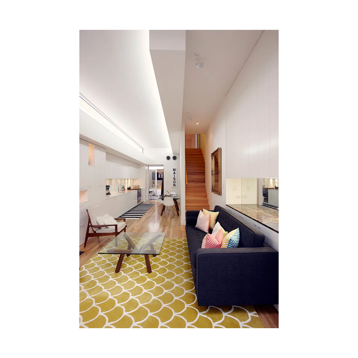 Very narrow white room with a navy sofa, yellow patterned rug, glass coffee table and a small dining table near the stairs.