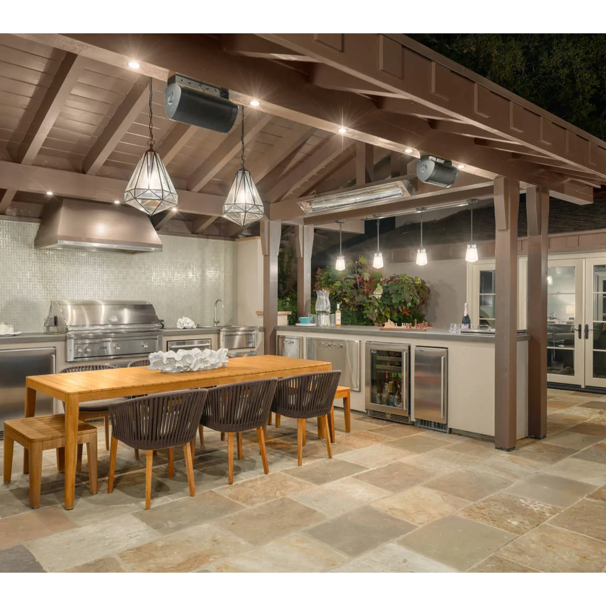 Covered outdoor kitchen with metal grill and hood, pale cabinetry, wood dining table and pendant lights