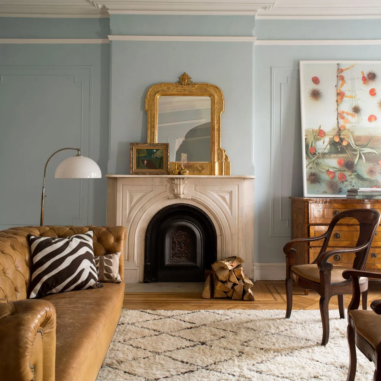 A brown tufted couch sits beside a pale fireplace, gold-framed mirror, wooden chair and patterned rug