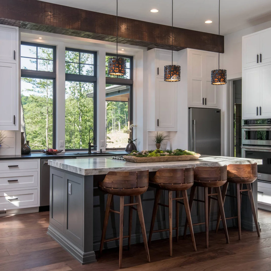 A gray island with wood stools beneath a dark beam in a white kitchen