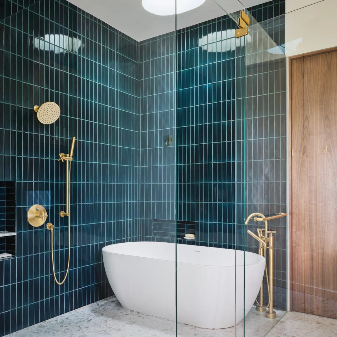 A white freestanding tub sits on pale terrazzo beneath deep blue vertical shower tiles.