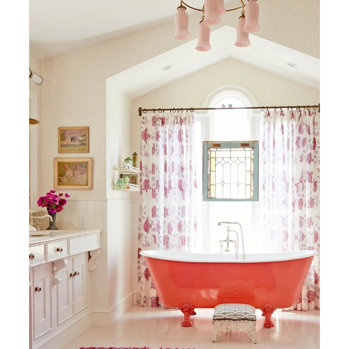 Pink floral curtains flank an arched window behind a coral-colored clawfoot bath and a suspended decorative glass panel.
