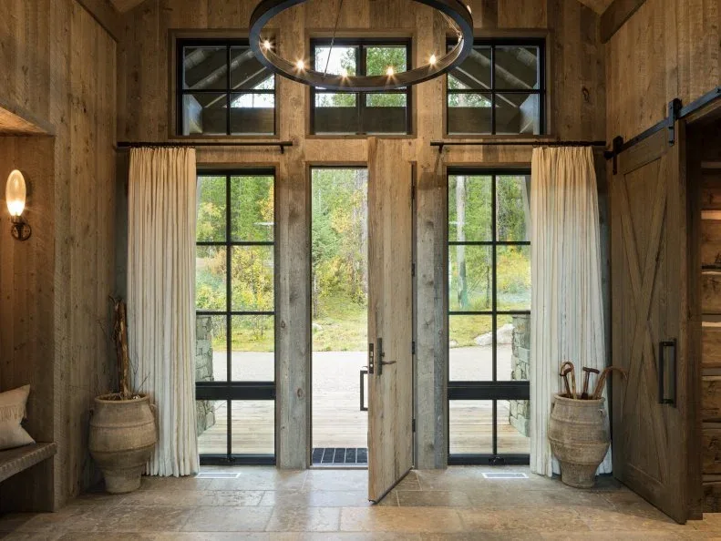 Dark circular chandelier with exposed lights in tall wood-paneled entry with glazed doors, upper windows, pale curtains and umbrella container
