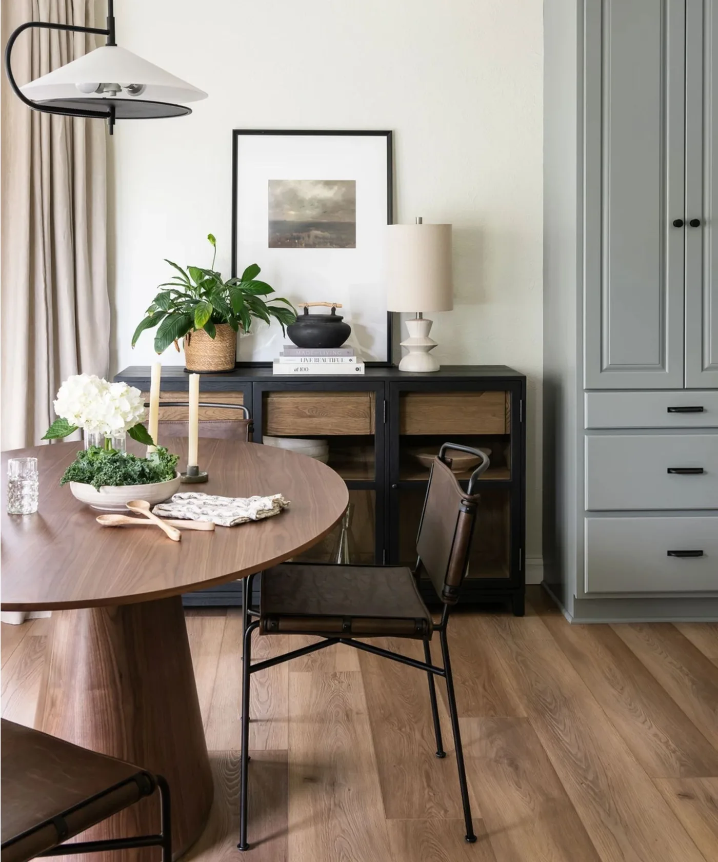 A black and wood-fronted dining sideboard stands behind dark-framed chairs and a round wood table beside pale curtains and a shaded table lamp