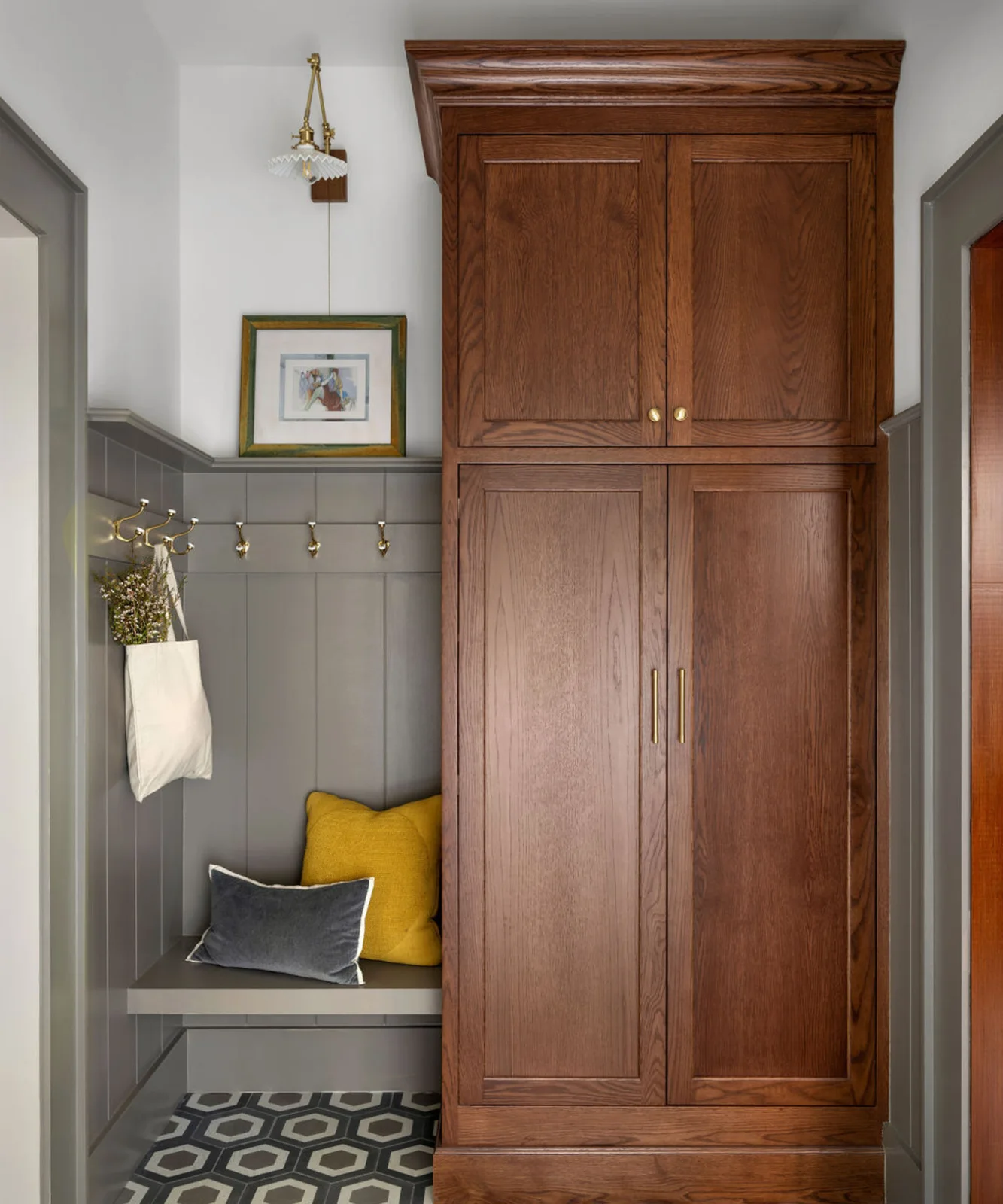 A tall wood mudroom cabinet stands beside a small gray bench, paneled hook wall, gold-colored fittings and a geometric floor.