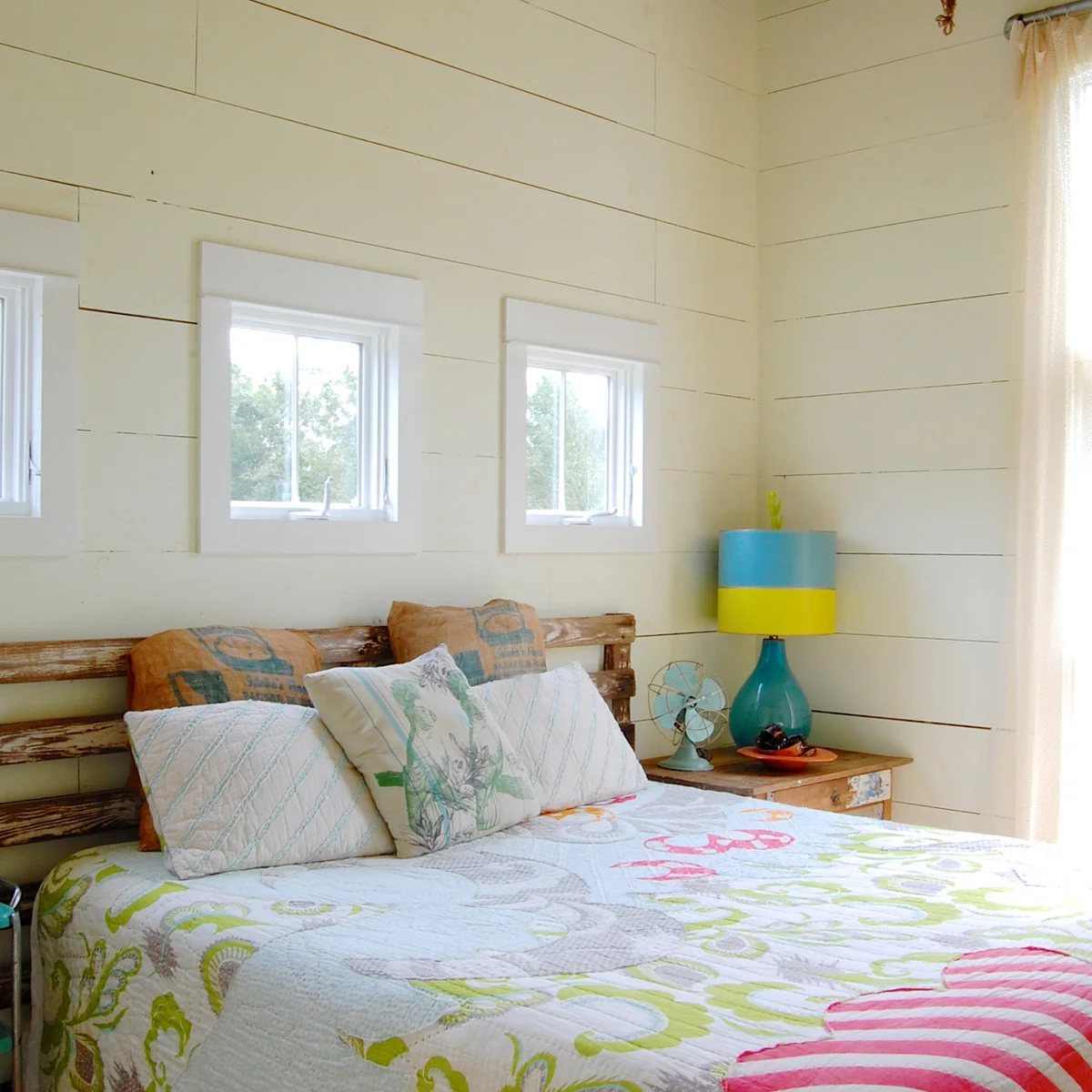 White plank bedroom with worn wood-colored headboard, colorful quilt and turquoise bedside lamp