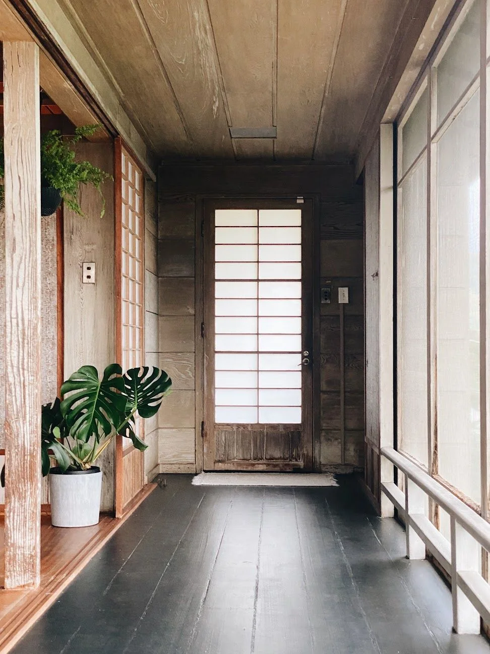A long low wooden bench runs beside translucent windows in an entrance with weathered wooden walls, dark board flooring and leafy plants