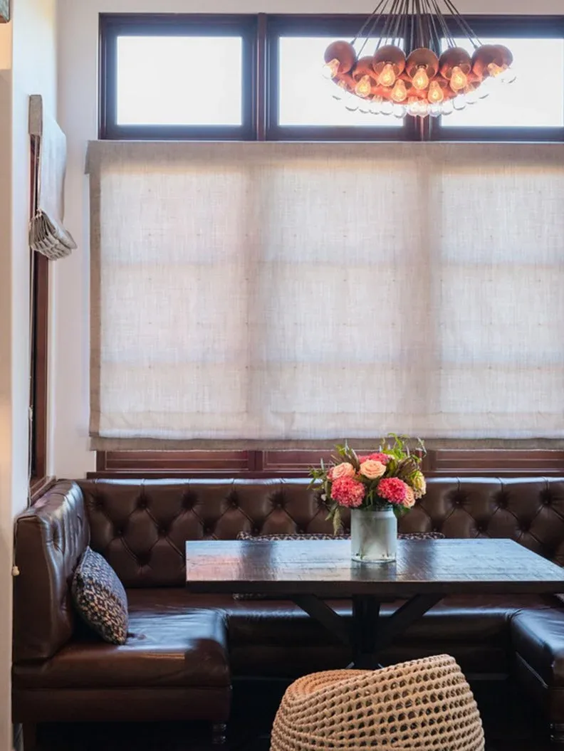 Dark brown tufted corner banquette beneath a broad window shade, with a dark rectangular table, patterned pillow, woven chair, flowers, and warm-colored overhead light.