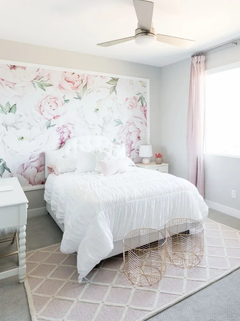 A framed pale pink floral mural sits behind a white bed with blush curtains and two open wire baskets.