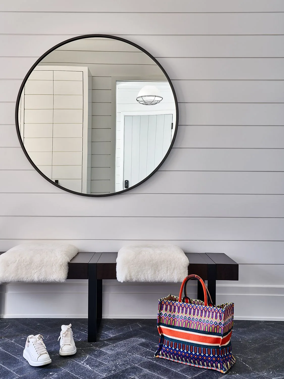 Dark simple entry bench beneath a large round mirror, with pale textured cushions, shoes and a striped woven bag