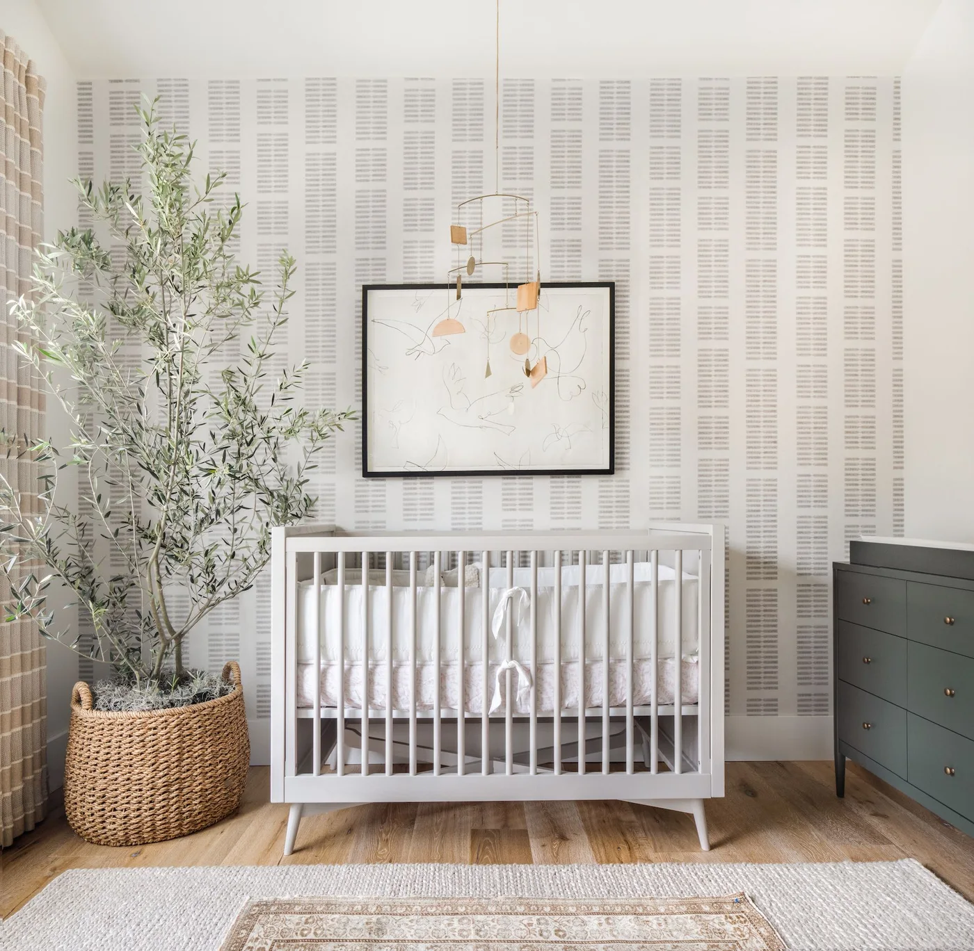 A pale nursery crib stands beside light gray repeated wallpaper, a dark gray dresser, a tall plant and a pale rug with a smaller patterned layer