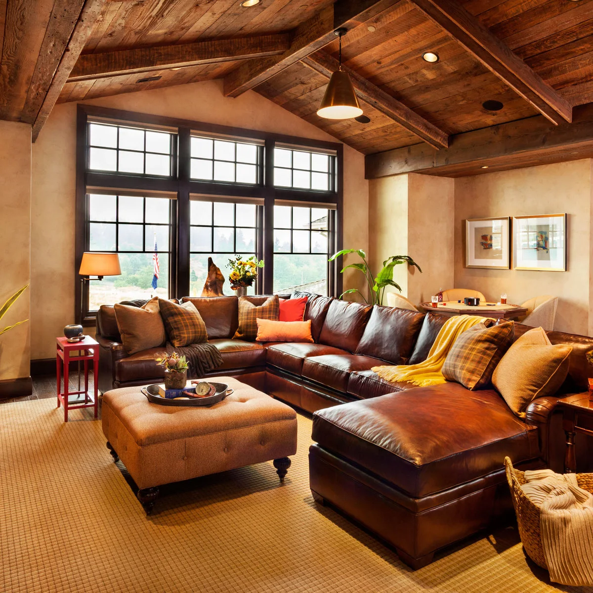 Dark brown leather sectional beneath a wood ceiling with rust cushions, a yellow throw and a large pale ottoman.