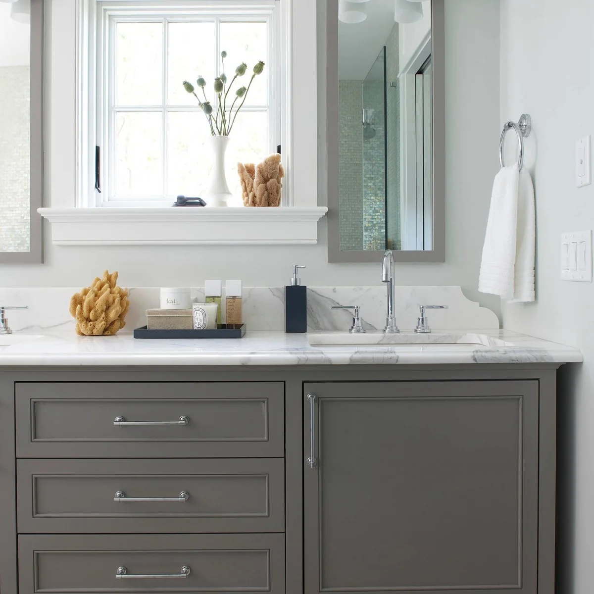 Gray double vanity with a dark tray of pale containers between two basins beneath a window.