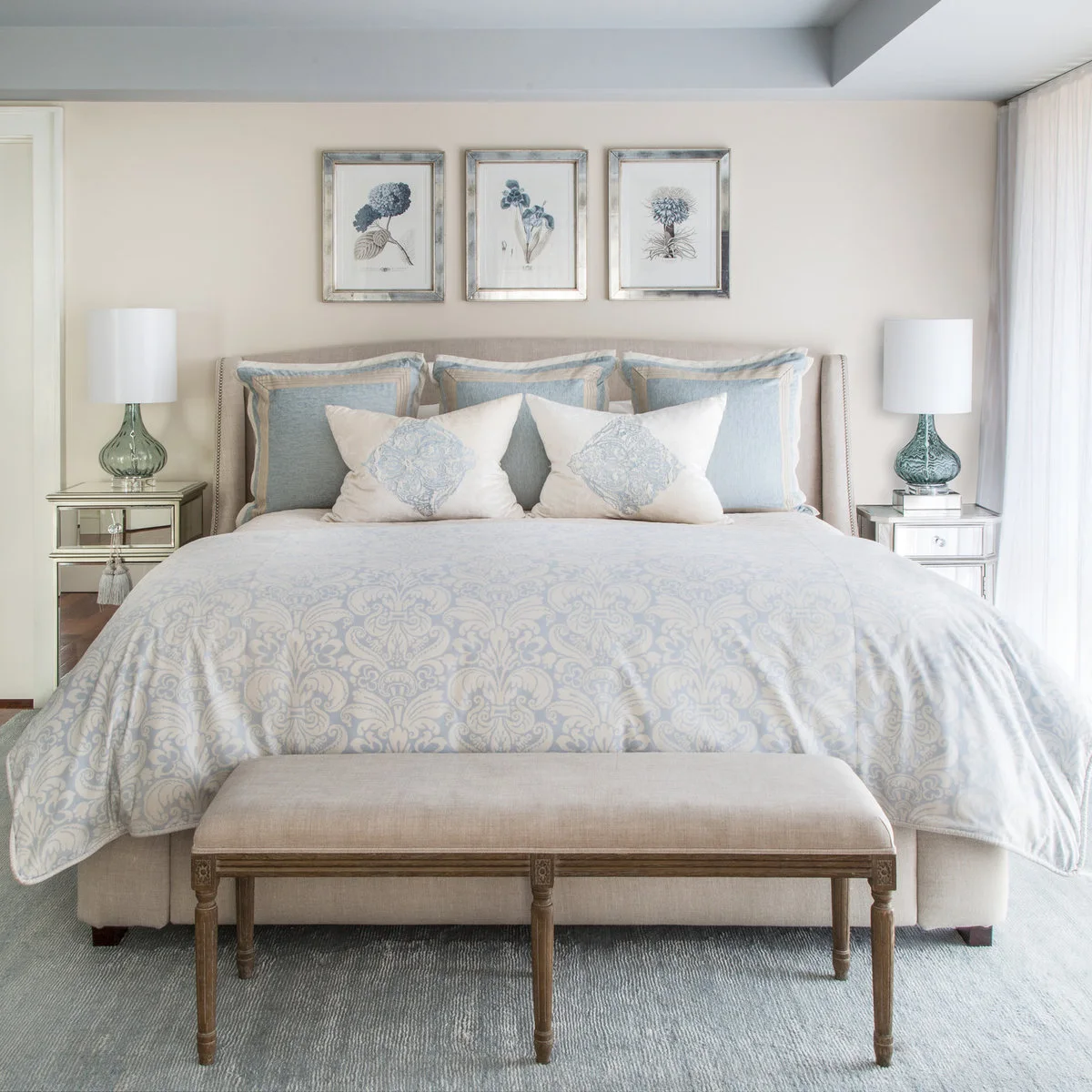 Bedroom with pale blue patterned bedding, a neutral upholstered bed, mirrored bedside cabinets and blue-green lamps
