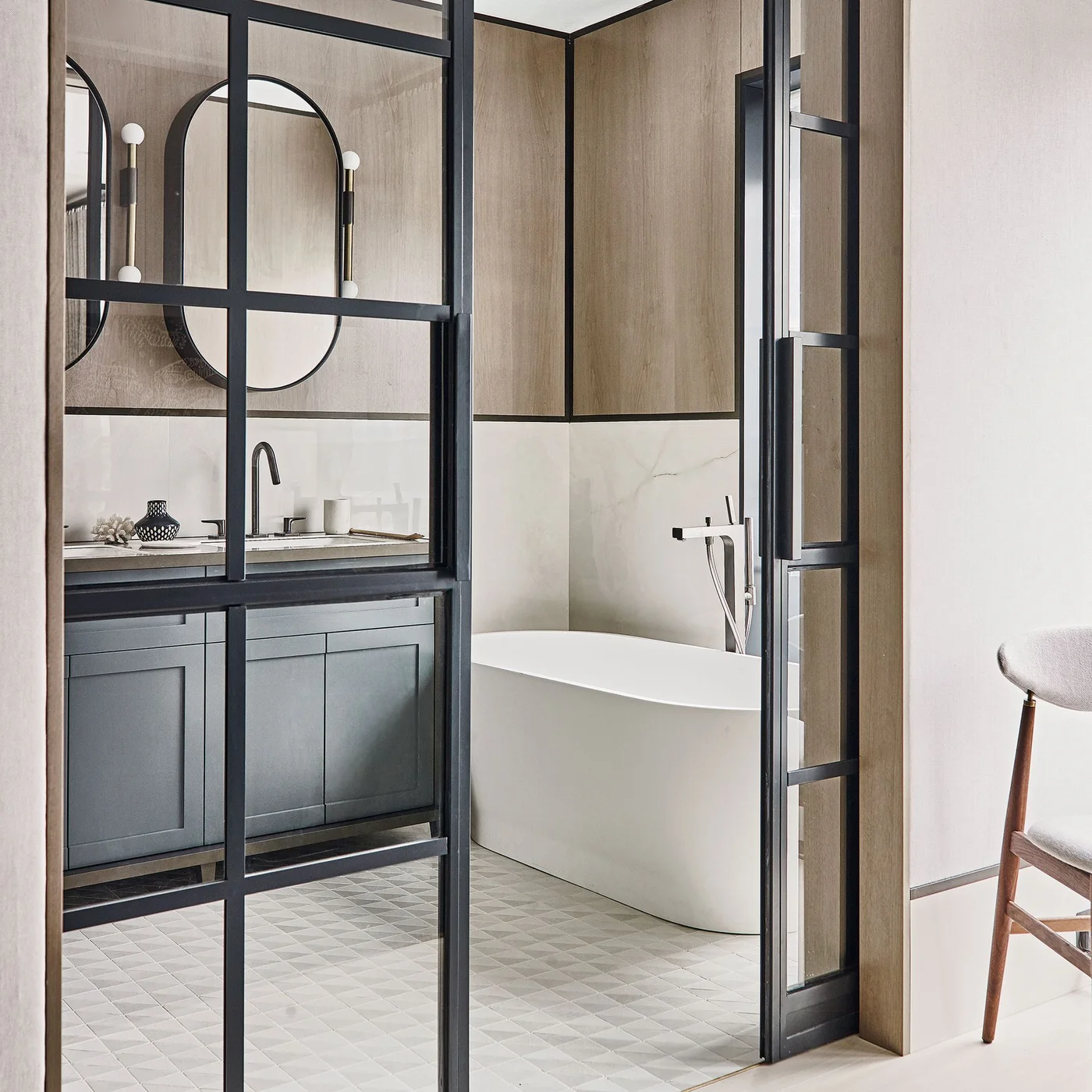 Black-framed glazed doors open toward a white freestanding bath and dark gray vanity with two long oval mirrors and white globe lights.