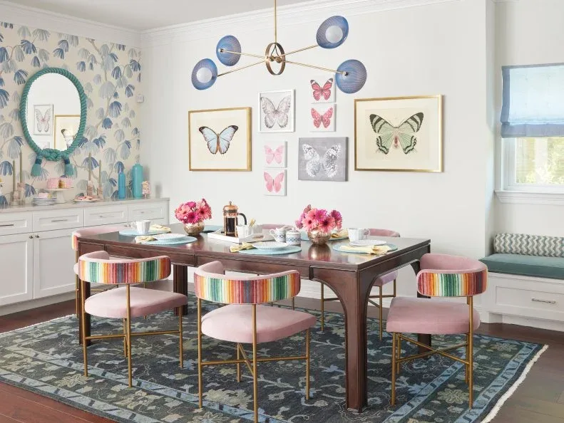 Mixed butterfly prints on a pale dining-room wall, with multicolored striped chair backs and pink seats, a dark wooden table, blue pendant shades, and a patterned adjoining wall.