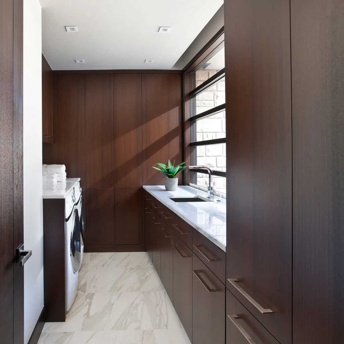 Narrow laundry room with dark wood cabinets, machines under a left counter and a sink beside the right window