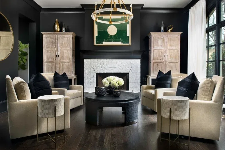 White brick fireplace beneath a black mantel in a dark room with pale chairs and paired wooden cabinets.