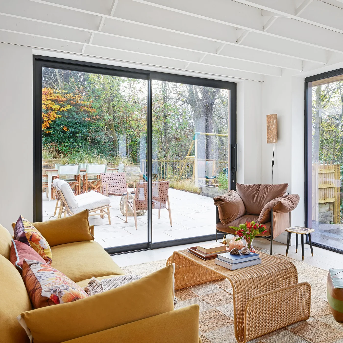 A yellow sofa and brown armchair sit around a woven table beside broad sliding garden doors.