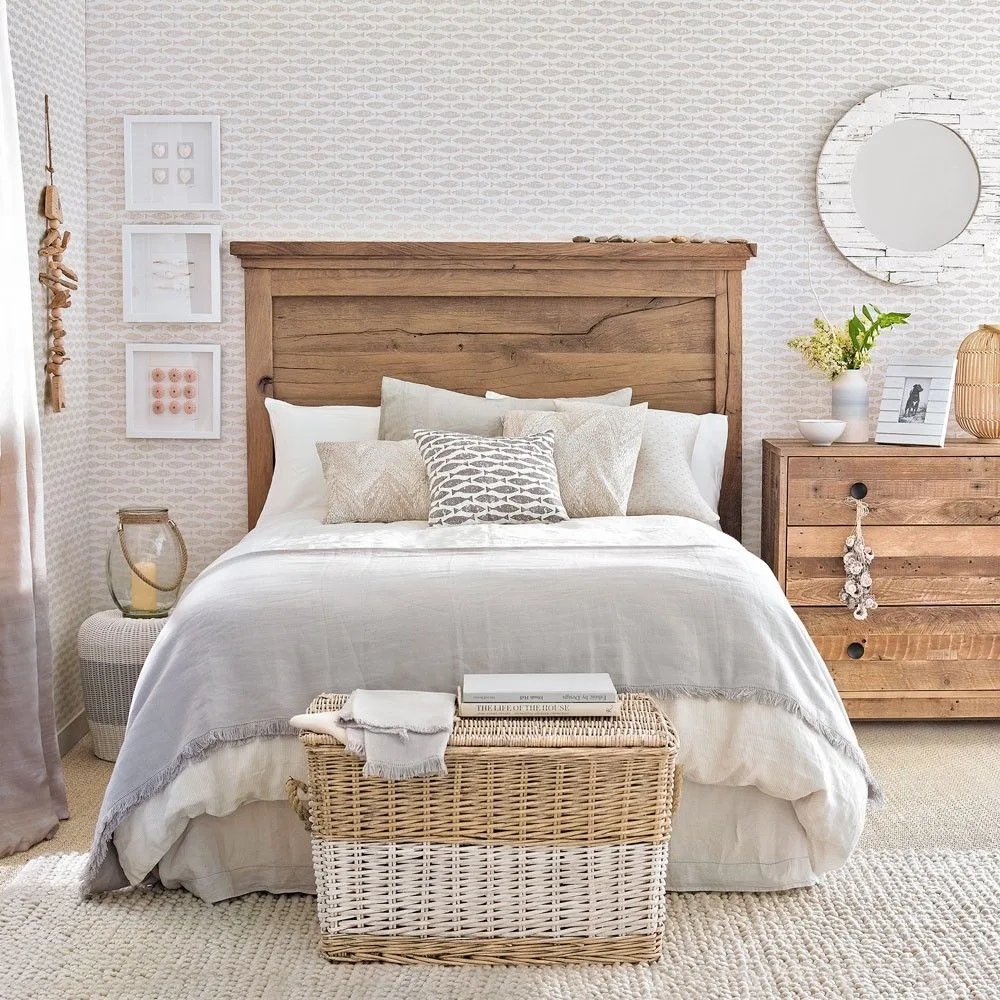 Neutral bedroom with small fish-pattern wallpaper, wooden bed and drawers, a round pale mirror and woven storage chest.