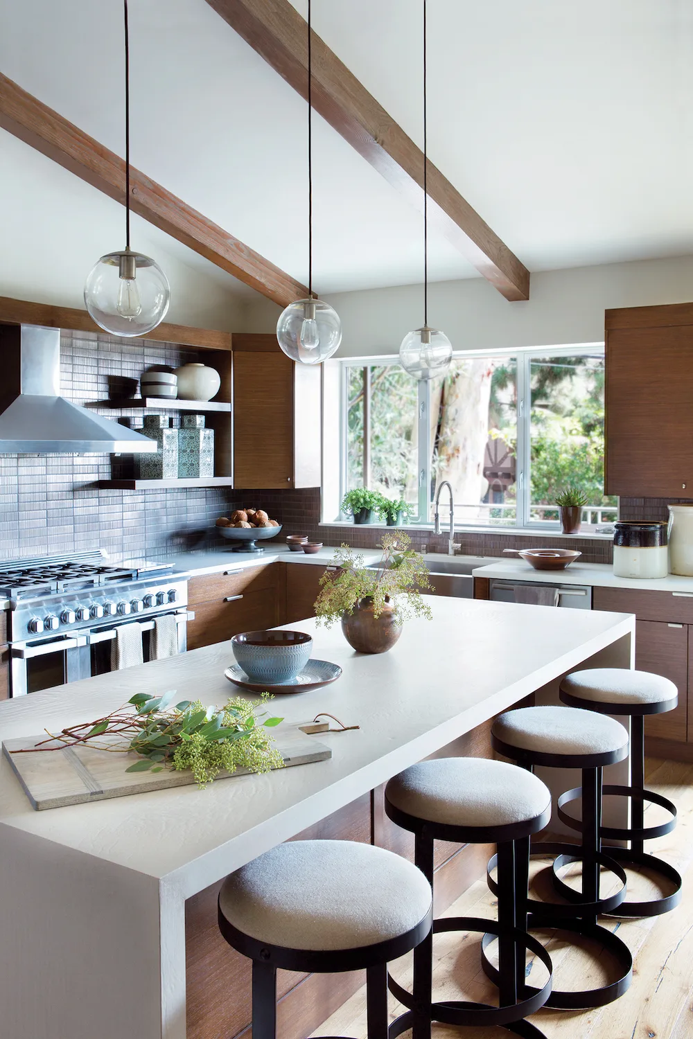 Kitchen with brown cabinets, broad sink window, white island counter, round pale stools, glass pendants, wood ceiling beams, and gray tile backsplash.