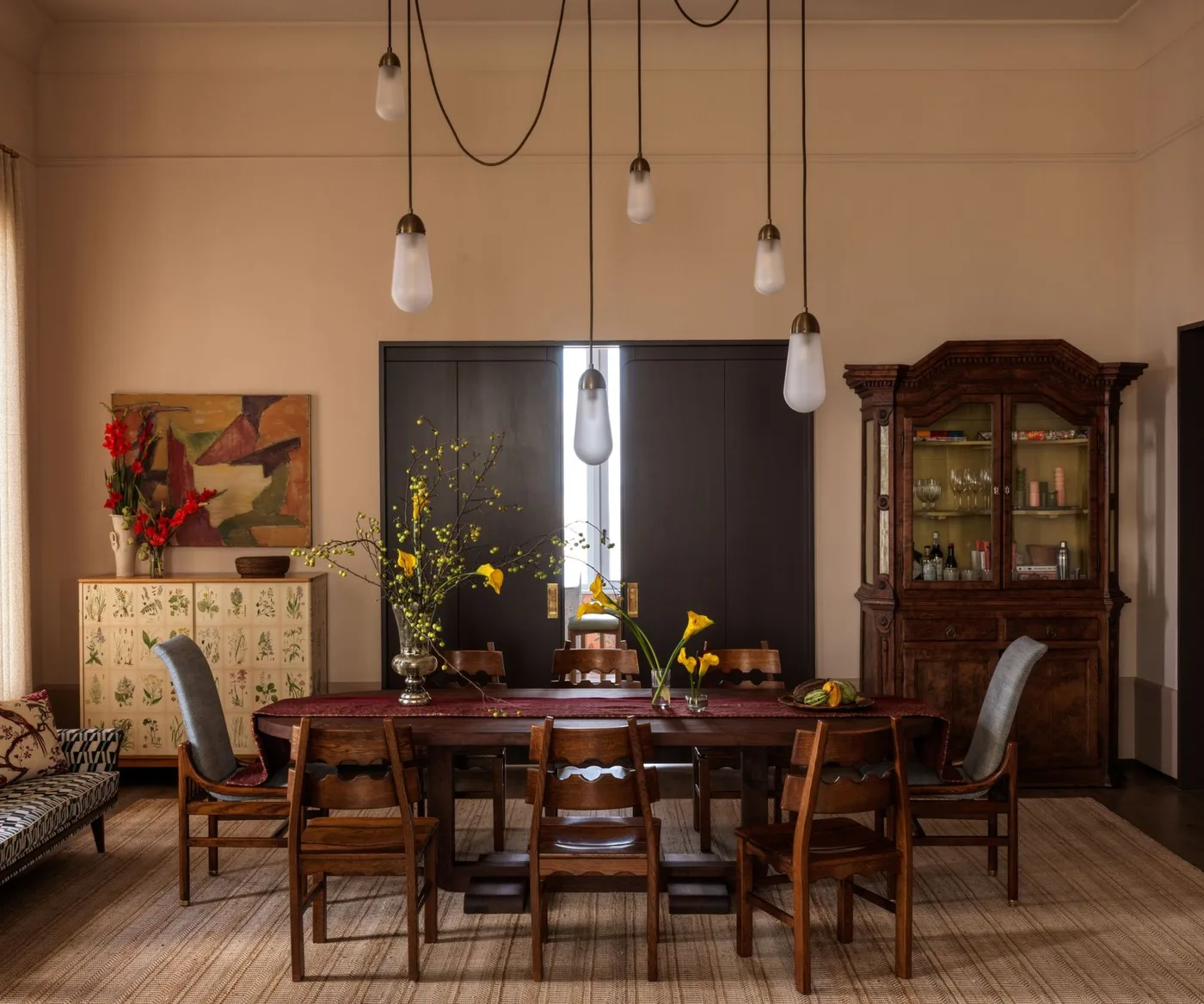 Country dining room with pale peach walls, wooden rectangular table, mixed chairs, dark glass-front cupboard and several hanging lights.