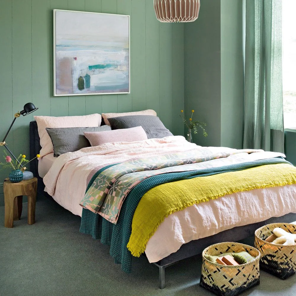 Green paneled walls and curtains frame pale pink bedding with gray cushions, layered green and yellow throws and woven baskets