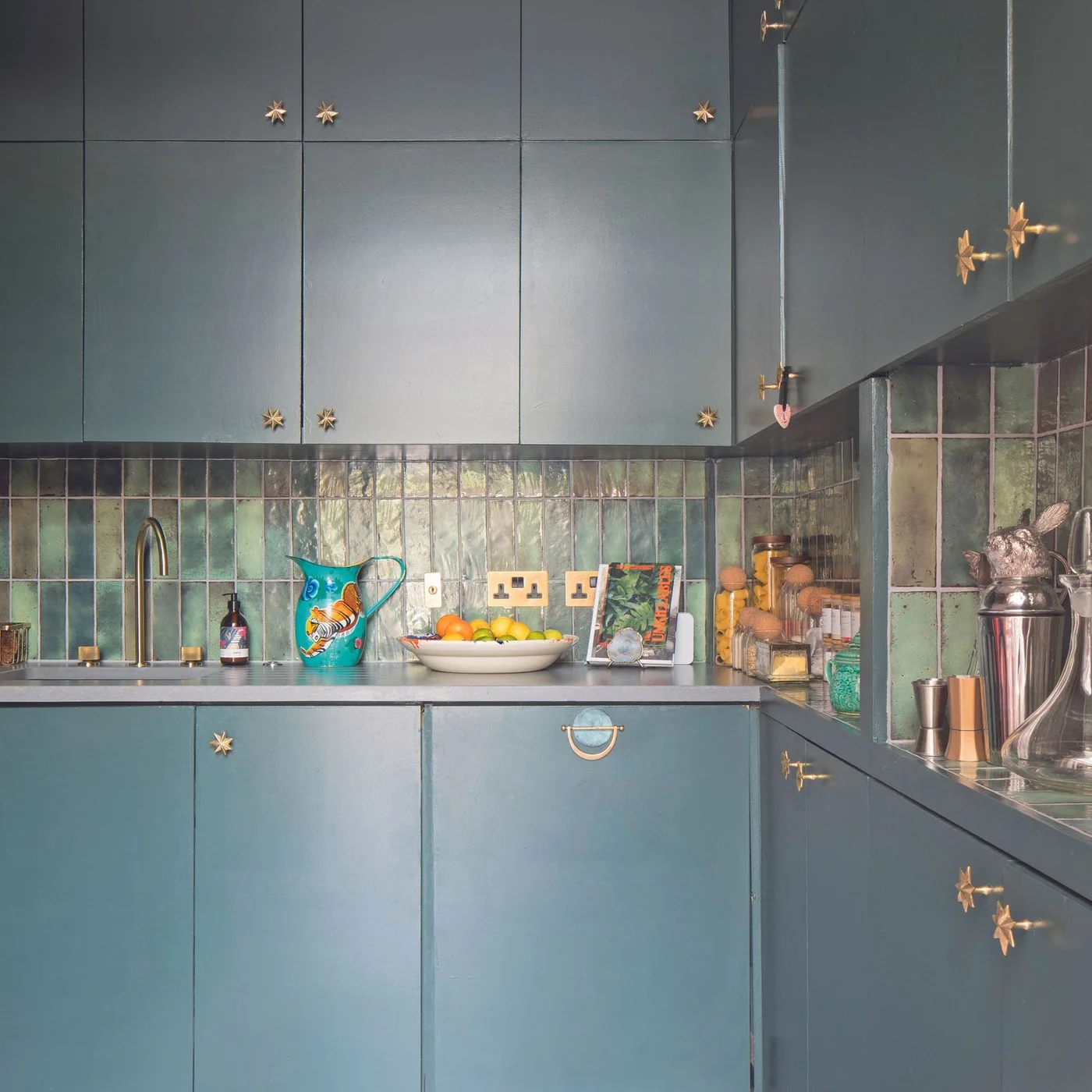 Deep teal cabinets with small warm-colored handles surround a glossy varied green tiled corner with a dark counter, jars and a colorful pitcher