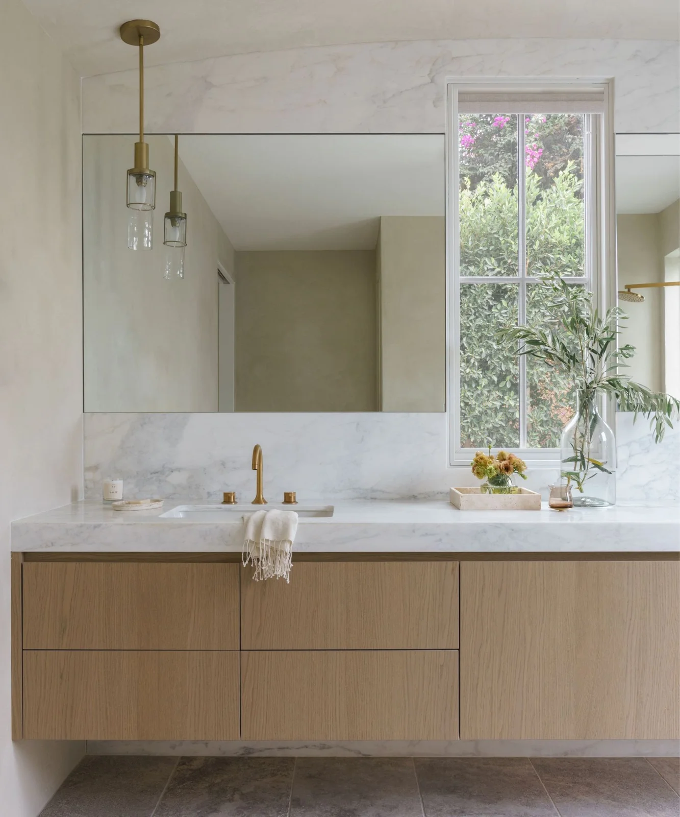 A floating wood-fronted vanity sits beneath a broad mirror beside a tall window, with pale veined surfaces and slender hanging lights