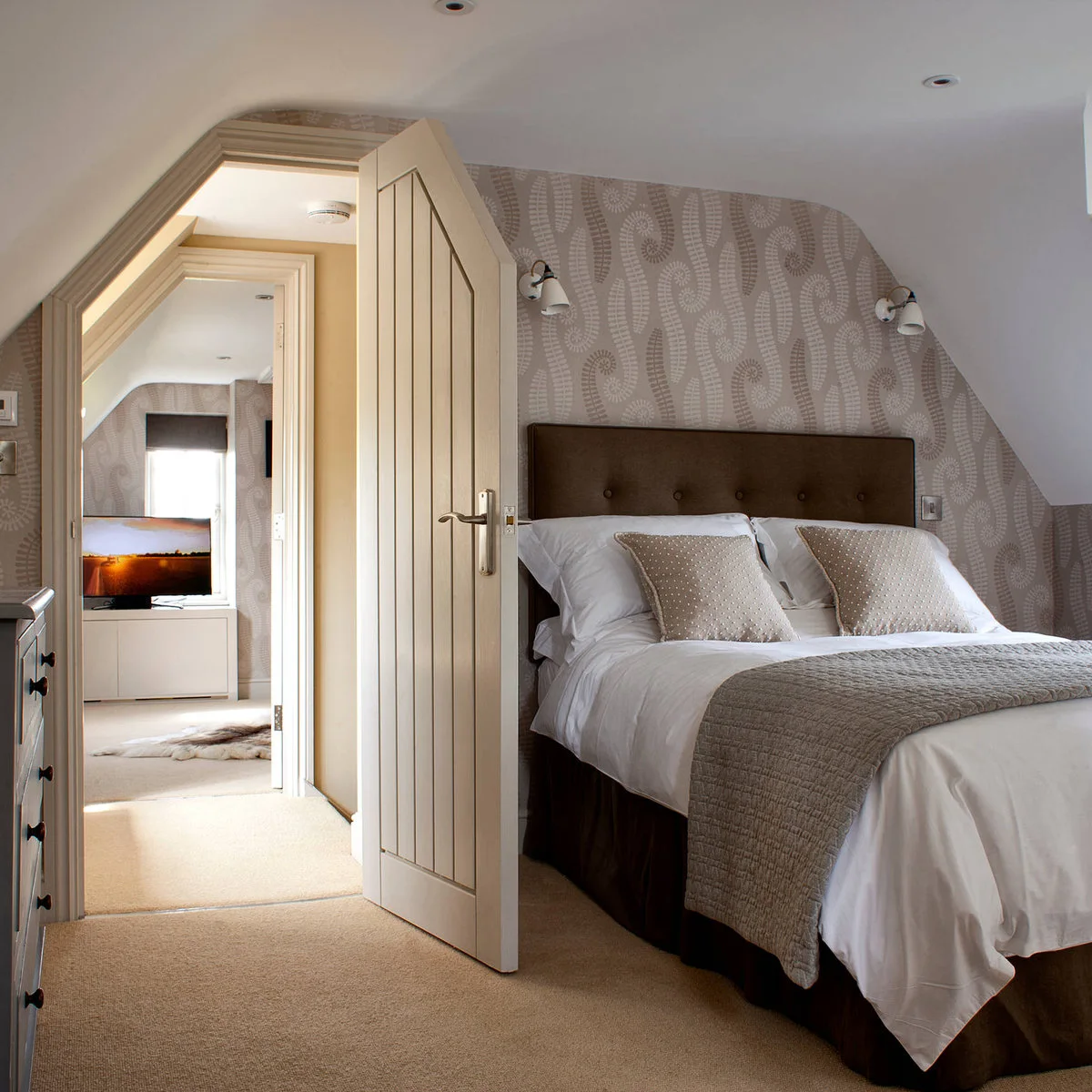 Low-ceilinged bedroom with patterned wallpaper, pale bedding and two small wall lights