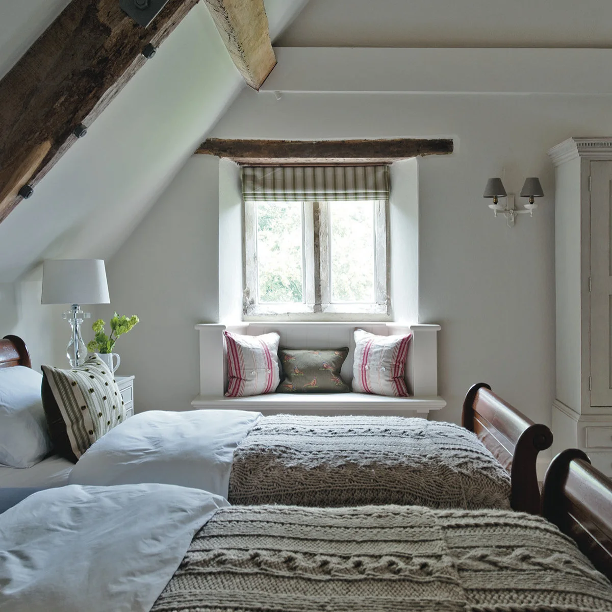 Twin wood-colored beds with gray knitted covers and a small window seat beyond