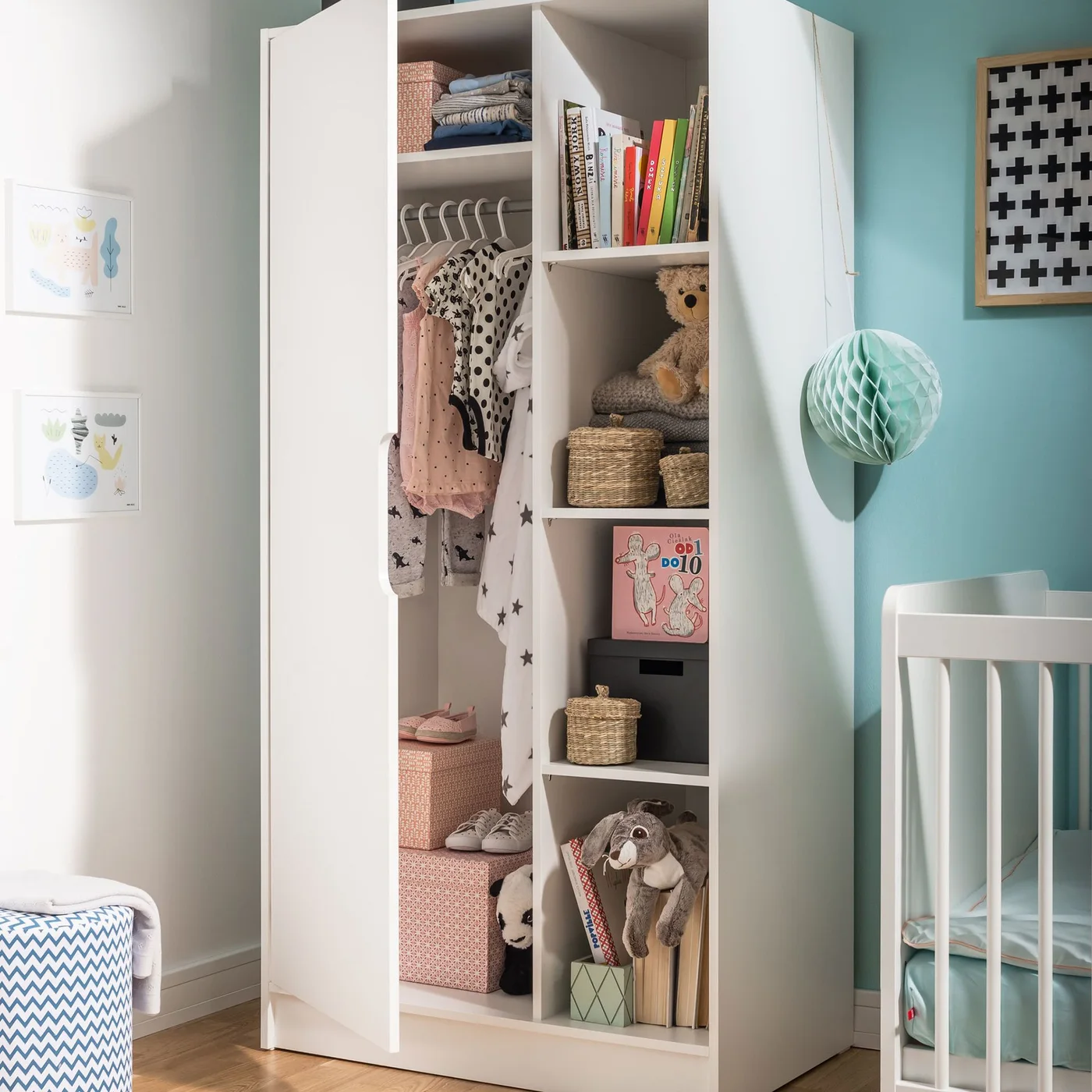 An open white nursery wardrobe has hanging clothes on one side and shelves with baskets, books and toys on the other.