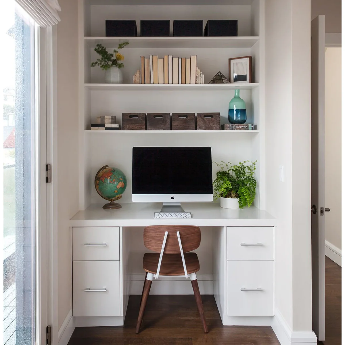 White alcove desk with a wood chair, two drawer units and open shelves beside a glass door
