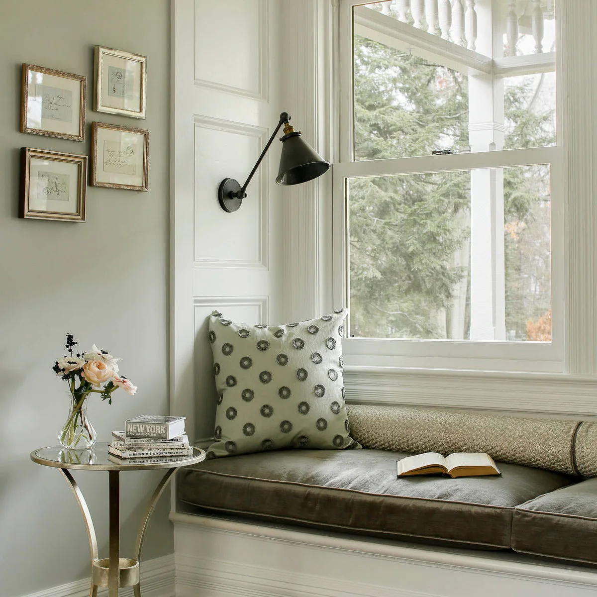 A dark upholstered window seat with a patterned pillow, adjustable wall lamp and small round table.