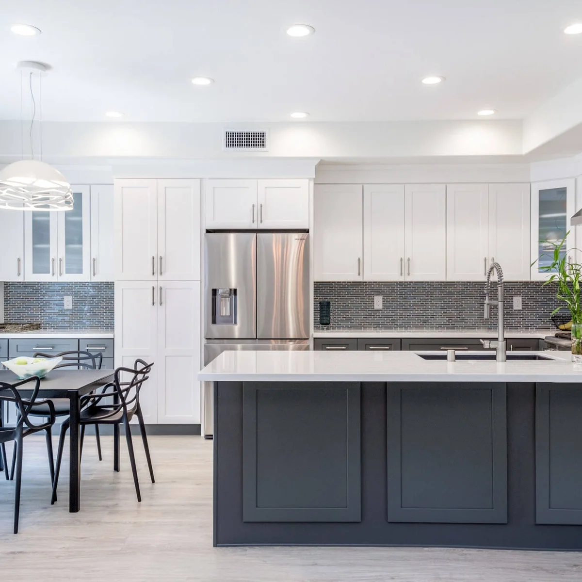 A charcoal peninsula and lower cabinets sit beneath white upper cupboards beside a narrow dark mosaic backsplash and black dining chairs.